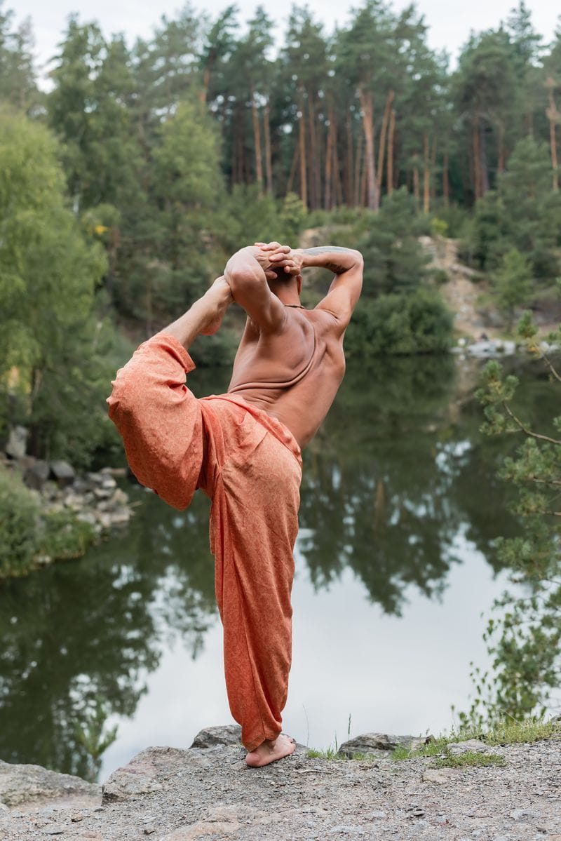 Thai fishermen pants on a shirtless Buddhist practicing one legged bow pose on rocky cliff over forest lake